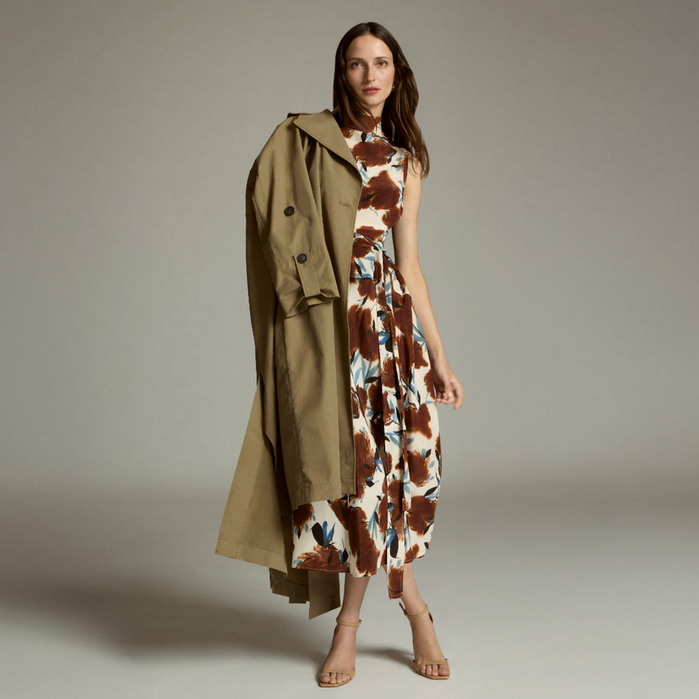 Woman wearing a patterned dress with a trench coat draped over her shoulder against a neutral background