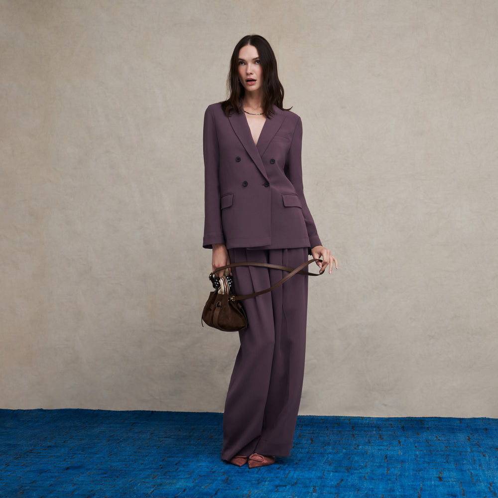 A model stands against a textured beige wall and blue floor, wearing a matching mauve double-breasted blazer and wide-leg trousers set, holding a brown handbag.