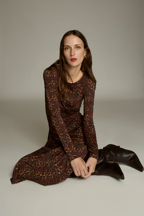 Woman in a brown patterned dress and black boots sitting on the floor against a plain background
