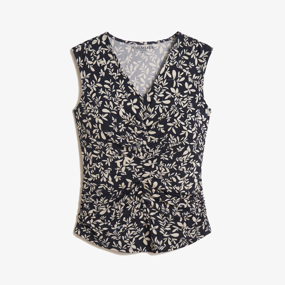 A sleeveless wrap-style top with an allover beige leaf and floral pattern on a dark navy background, featuring a V-neckline and gathered waist details, shown against a light gray background.