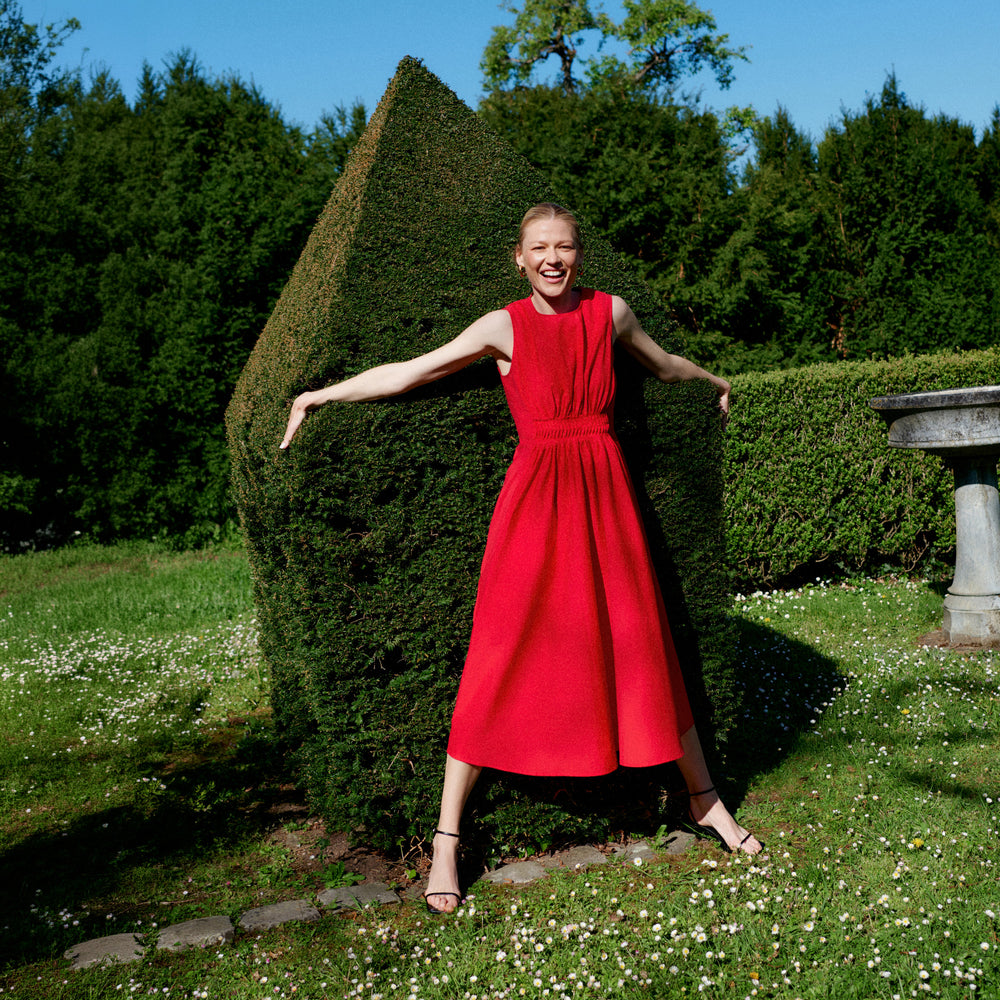 Woman in a red dress standing in a garden with greenery and a clear sky.