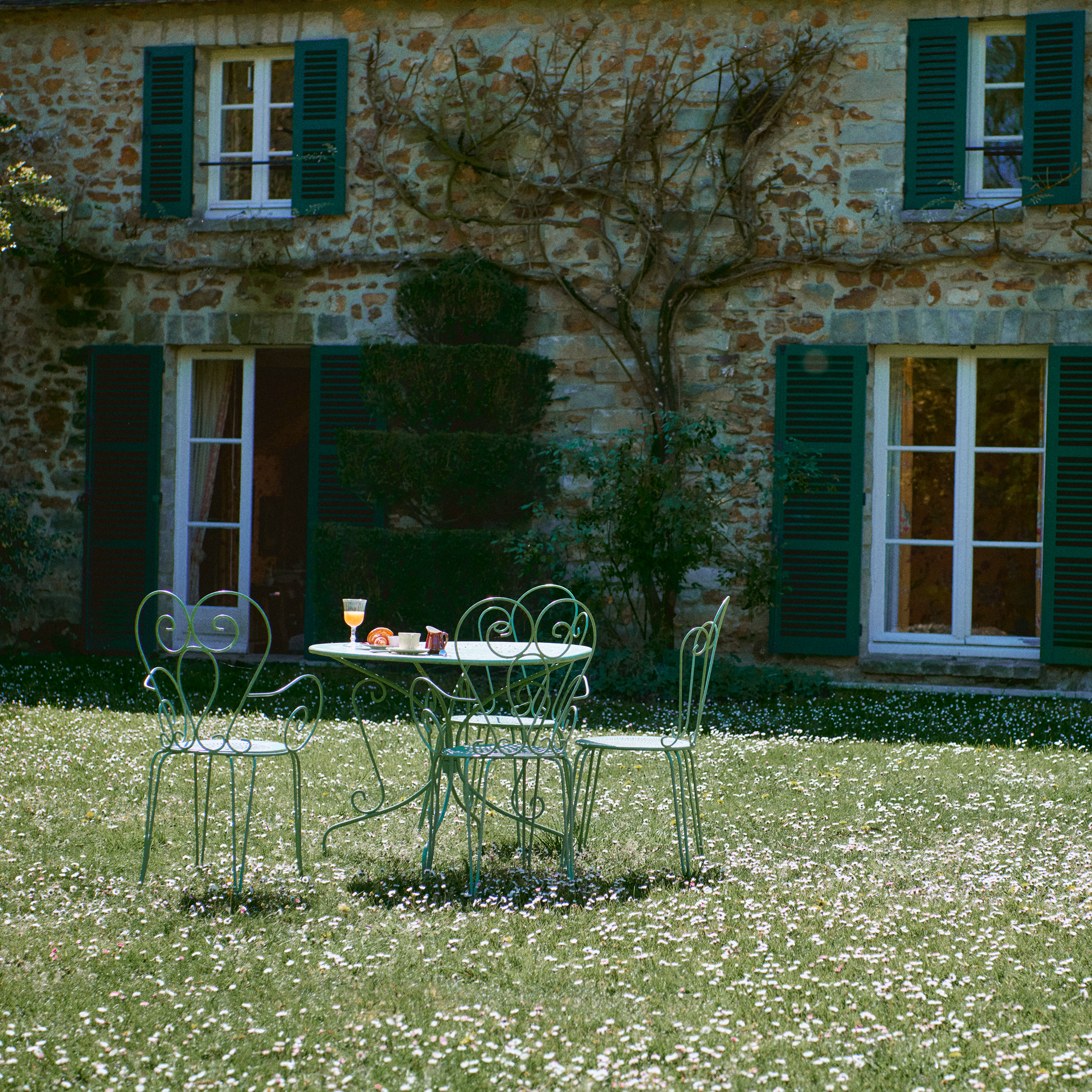 Outdoor setting with a stone building, green chairs, and a table on a grassy area.