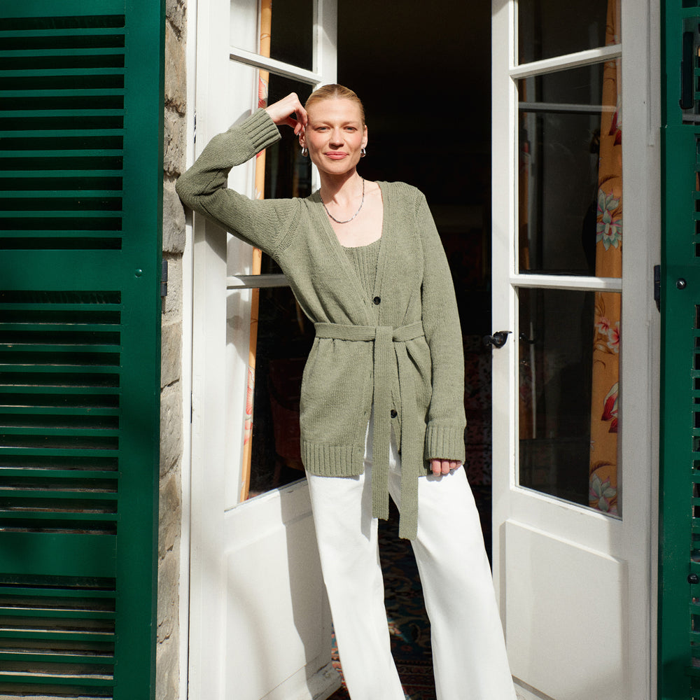 Woman in a green cardigan standing in an open doorway with green shutters.