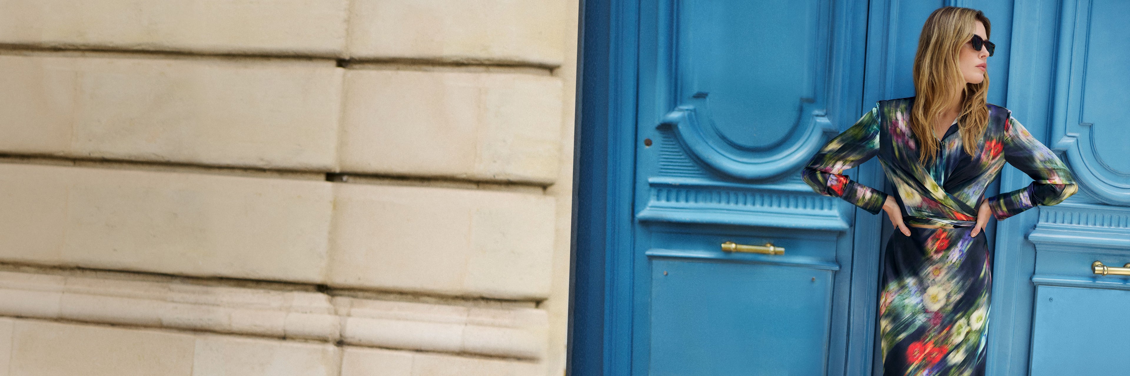 A woman wearing a colorful, long-sleeved abstract print dress and sunglasses stands in front of a bright blue door, with a beige stone wall on the left.