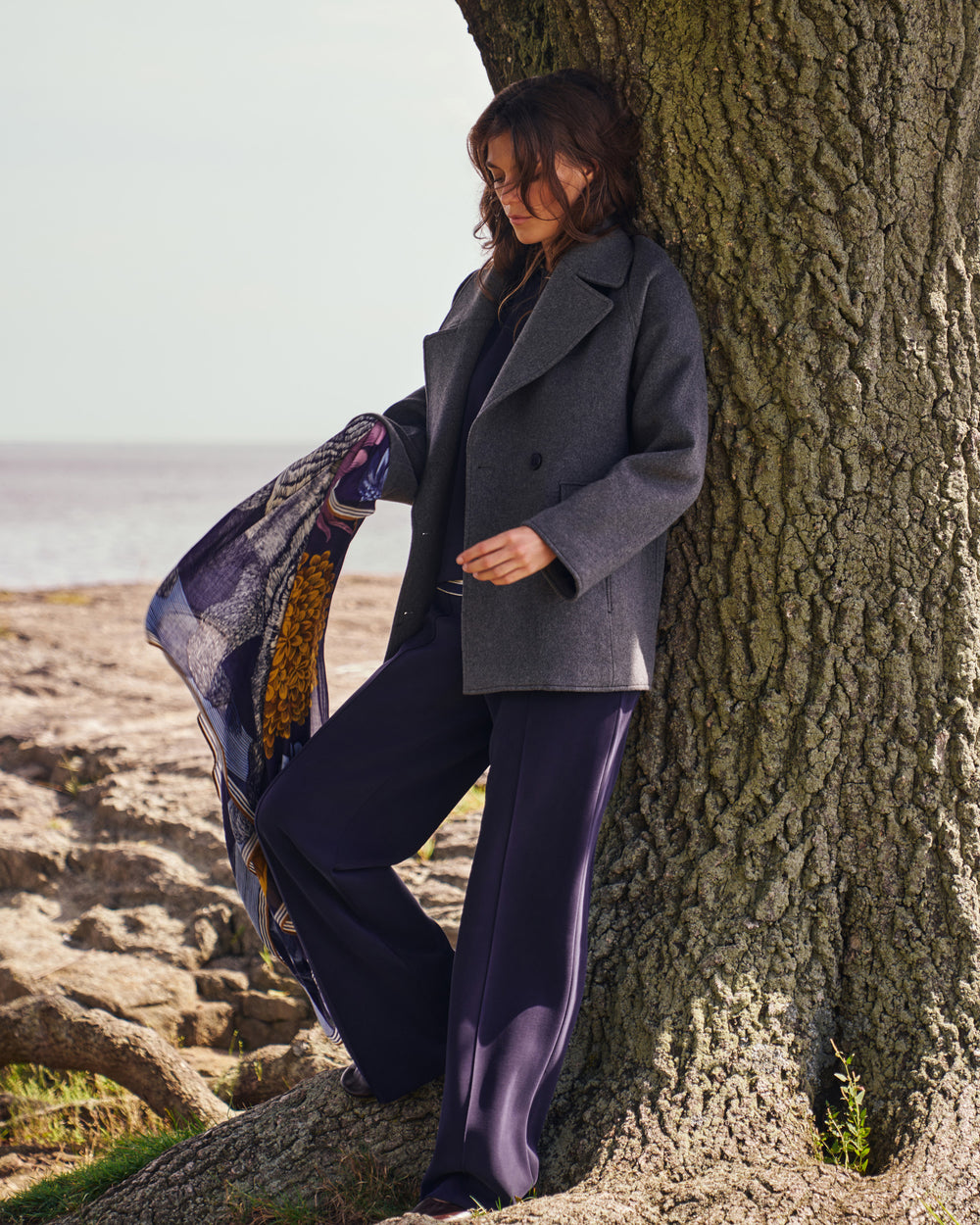 A woman leans against a large tree outdoors near the shore, wearing a charcoal gray double-breasted coat, dark purple wide-leg trousers, and holding a patterned scarf.
