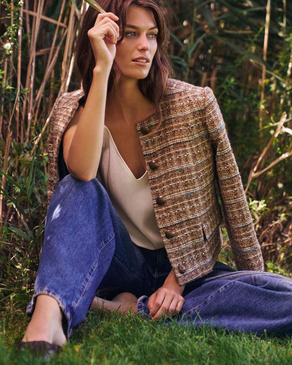 A woman sits on grass outdoors wearing a brown tweed blazer over a cream camisole and blue jeans, with tall reeds and greenery in the background.