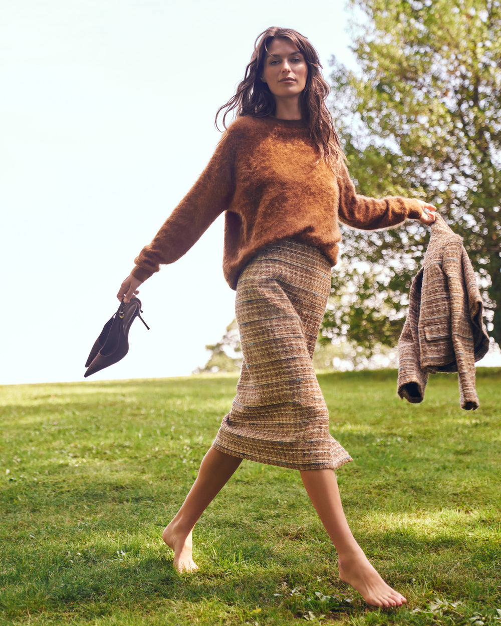 A woman walking barefoot on a grassy lawn wearing a fuzzy brown sweater, a brown and beige plaid midi skirt, and holding a matching plaid jacket and black pointed shoes.
