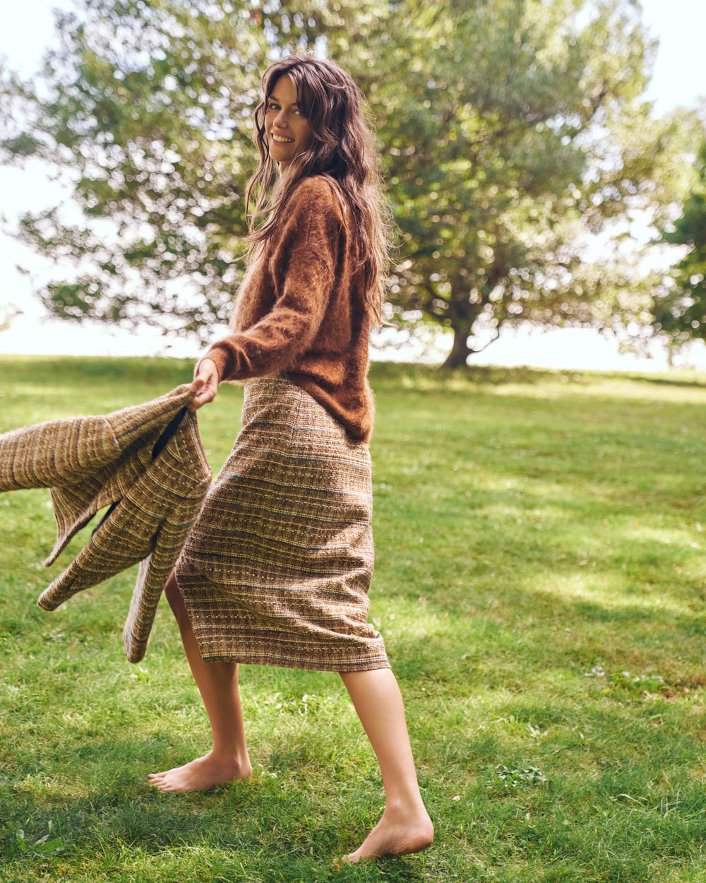 A woman stands barefoot on a grassy lawn outdoors, holding a plaid blanket or throw in one hand while wearing a brown fuzzy sweater and a brown-and-beige plaid midi skirt. Trees and bright sky are blurred in the background.