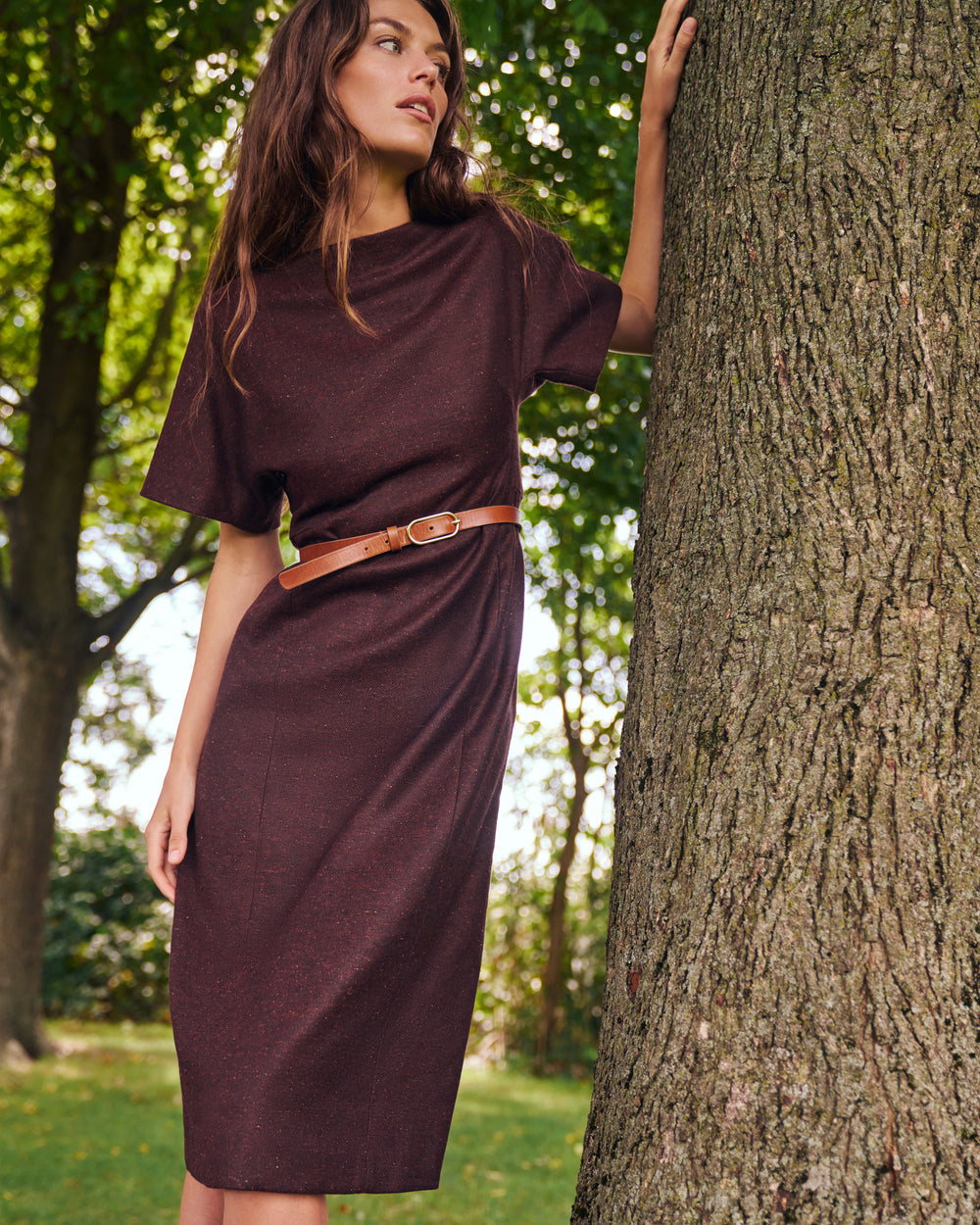 A woman wearing a short-sleeve, knee-length dark plum dress with a brown belt, standing outdoors next to a large tree trunk.