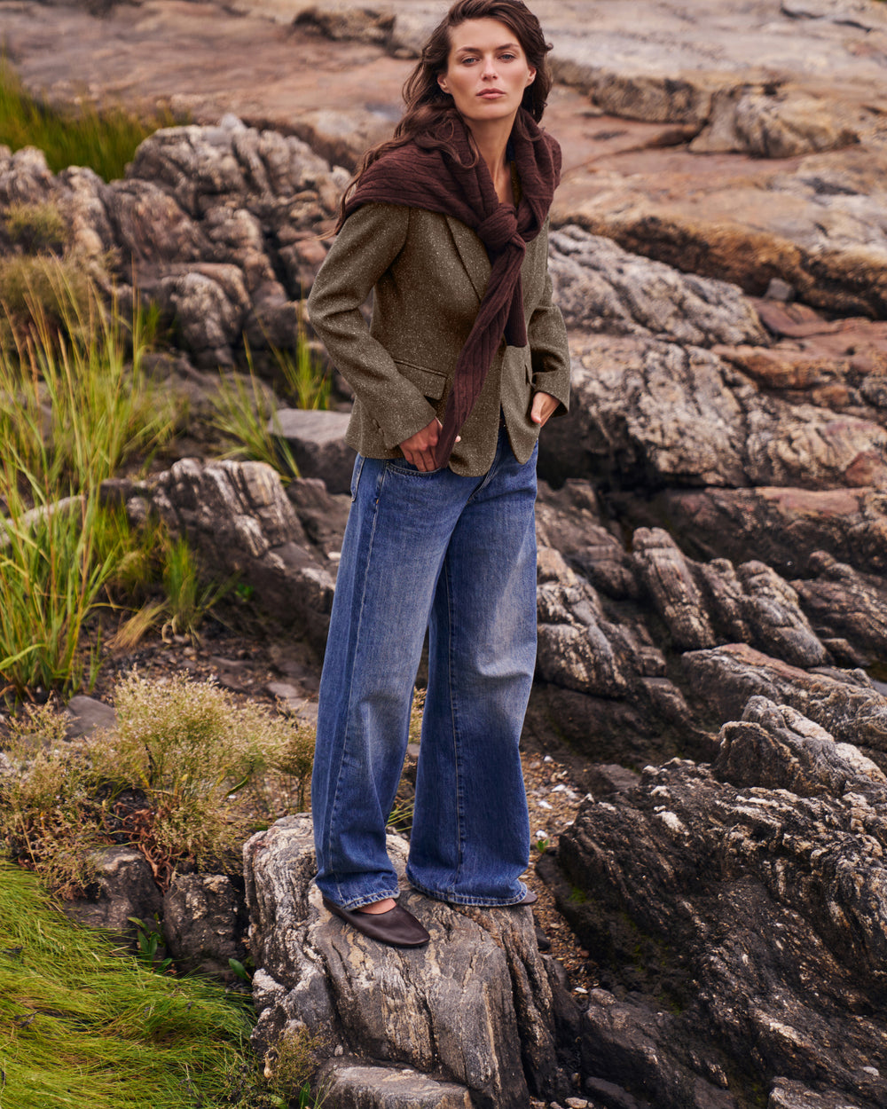 A woman stands on a rocky outdoor shoreline wearing a textured olive green jacket, wide-leg blue jeans, dark brown shoes, and a dark brown scarf.
