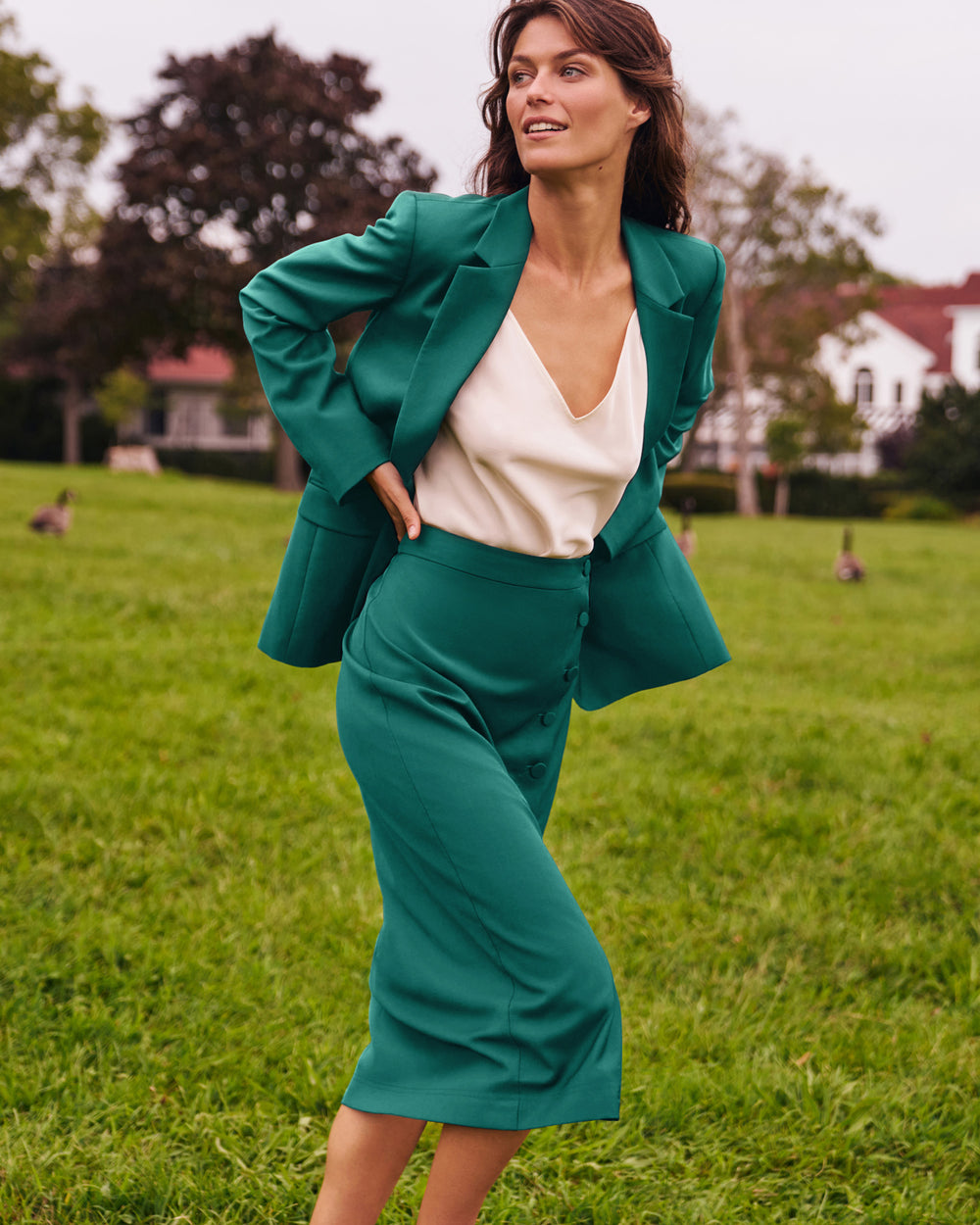 A woman posing outdoors in a green two-piece suit consisting of a blazer and matching midi skirt, layered over a cream V-neck top, set against a blurred green lawn background.