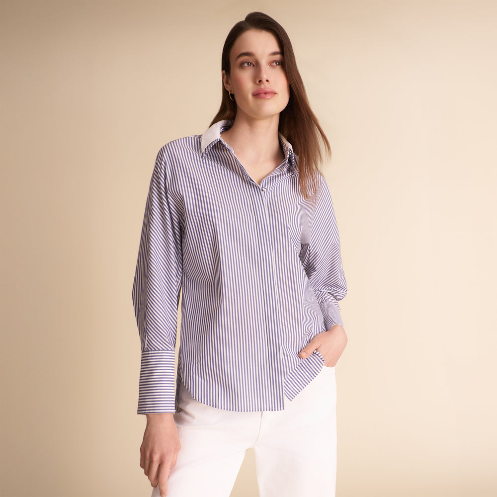 A woman modeling a long-sleeve button-up shirt with thin vertical blue and white stripes, paired with white pants, against a solid beige studio background.