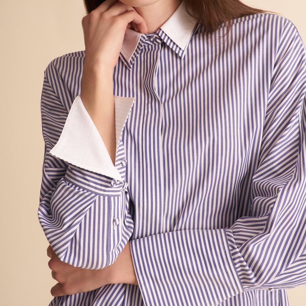 A close-up of a woman wearing an oversized long-sleeve button-up shirt with vertical blue and white stripes, featuring a solid white pointed collar and turned-back cuffs.