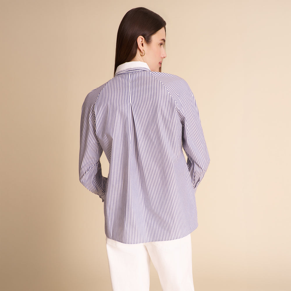 A woman shown from the back wearing a lavender and white vertically striped button-down shirt with white trousers, set against a solid beige studio background.