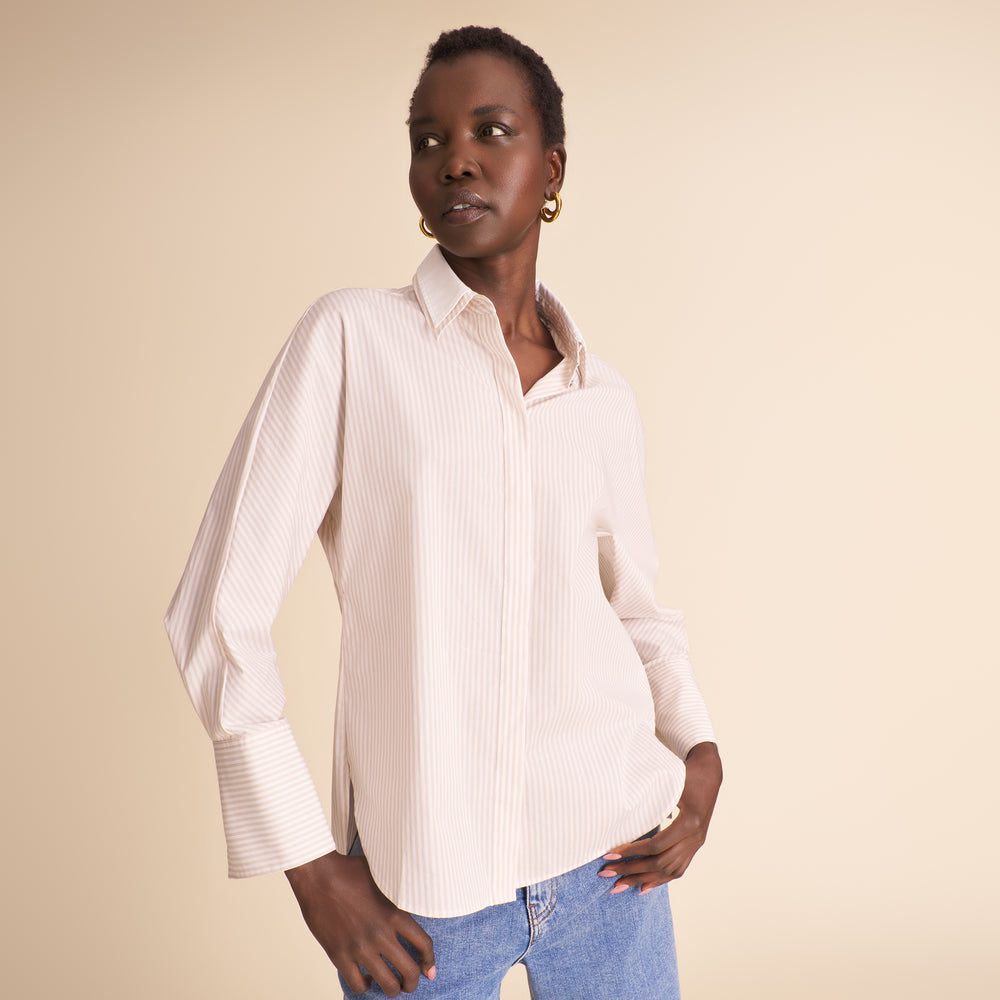 A woman modeling a light beige and white vertically striped button-down shirt with blue jeans, set against a solid beige studio background.