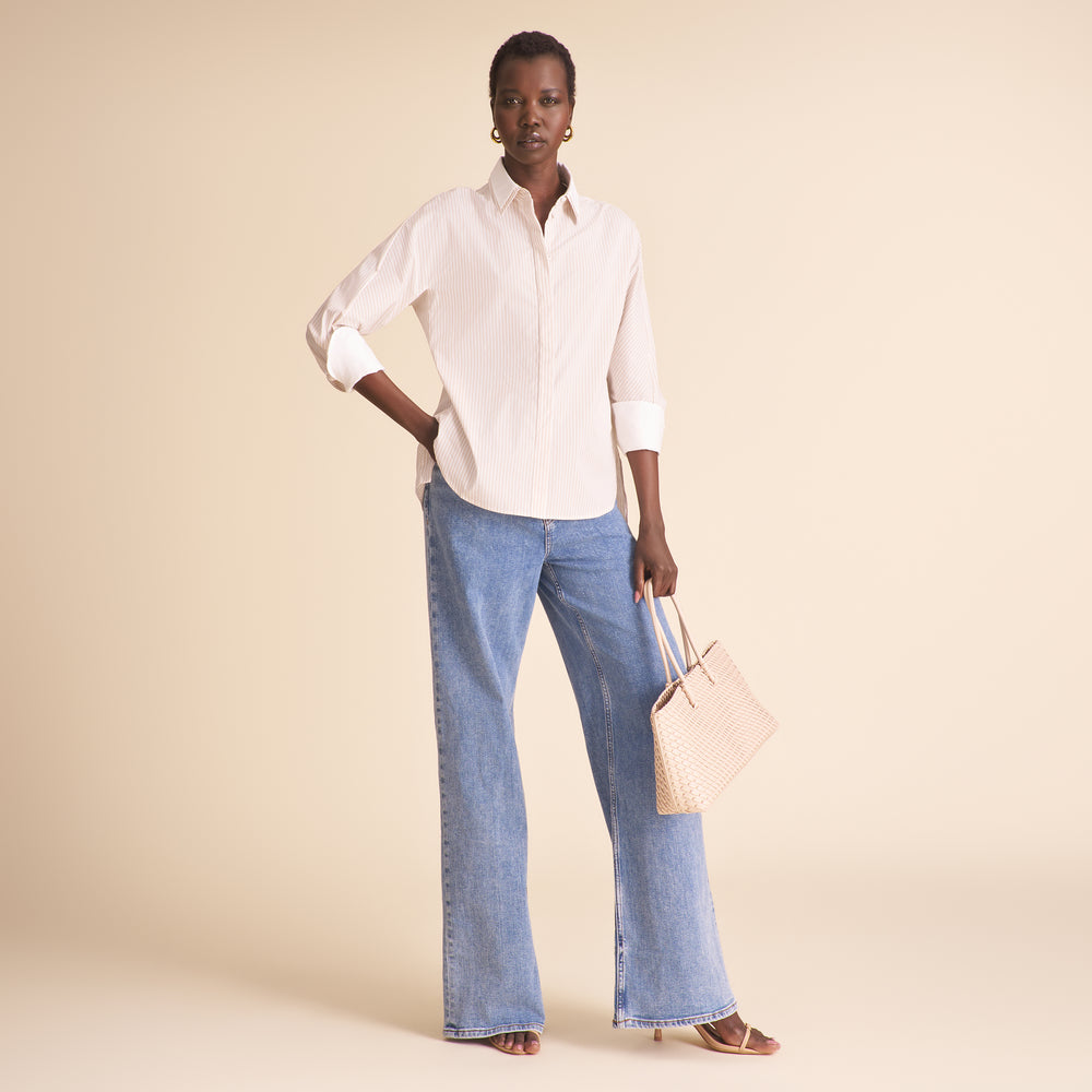 A model stands against a solid beige studio background wearing a white long-sleeve button-down shirt, light-wash wide-leg blue jeans, and holding a light pink woven tote bag.