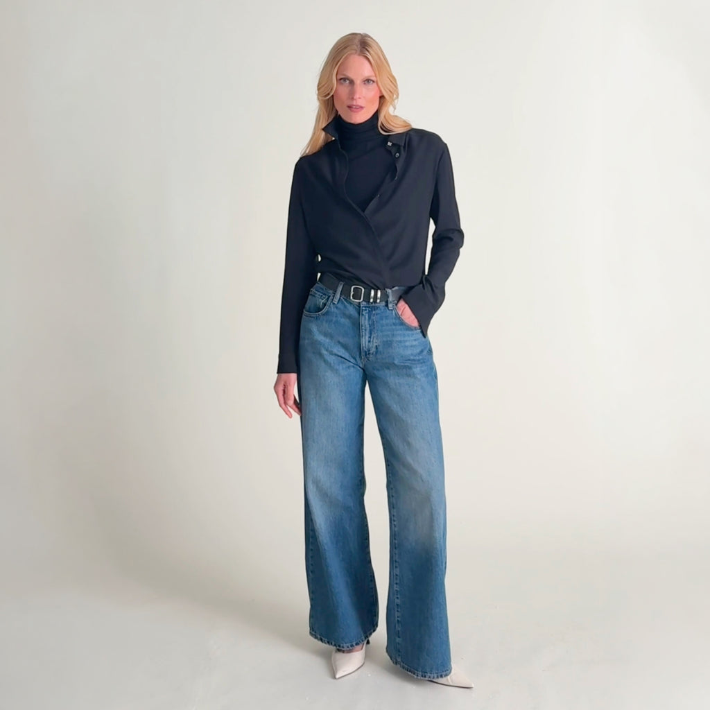 Video of a woman wearing a black long-sleeve button-up shirt tucked into blue jeans with a black belt. 