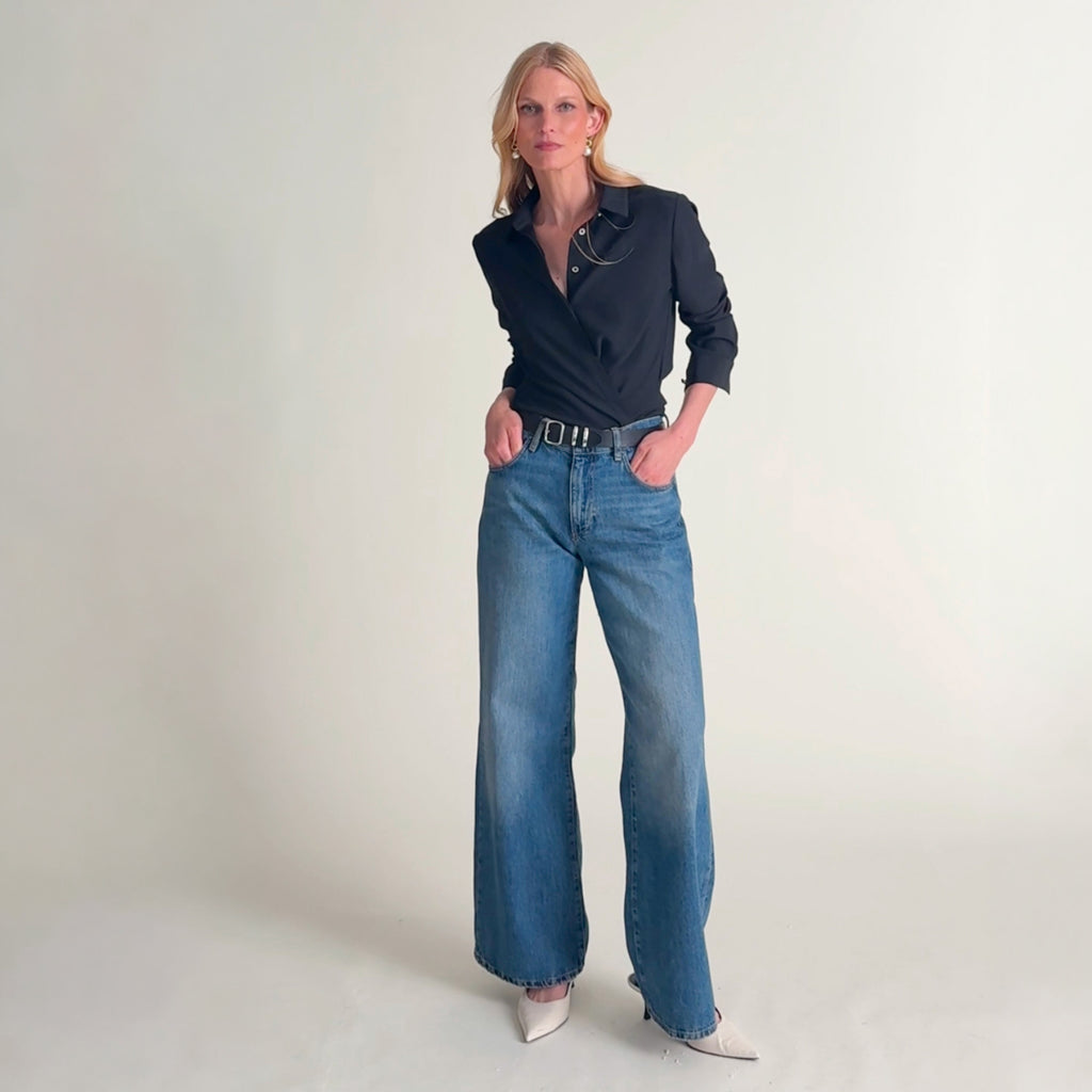 Video of a woman modeling a black long-sleeve button-up shirt and wide-leg blue jeans against a plain light beige studio background.