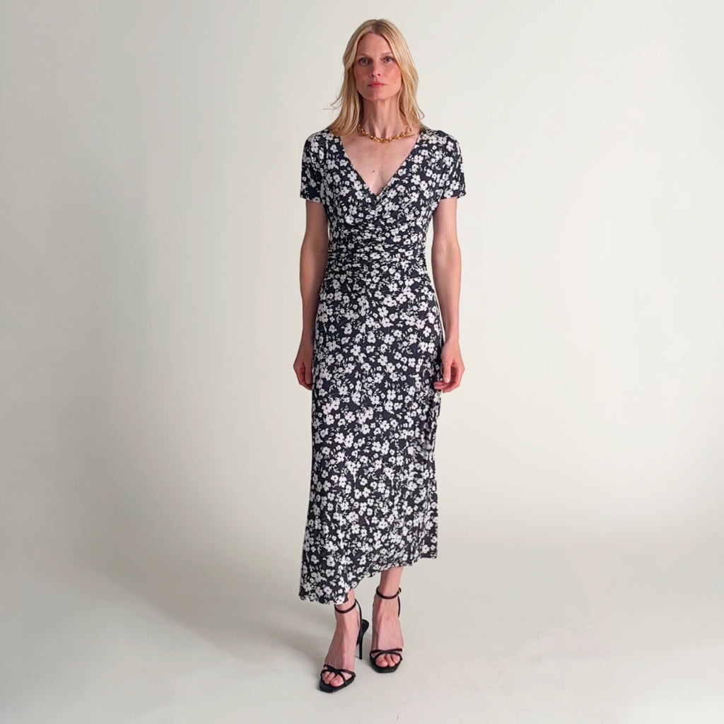 Video of a woman wearing a short-sleeve wrap-style dress with a dark navy background and an all-over white floral print, photographed against a plain light beige studio background. 